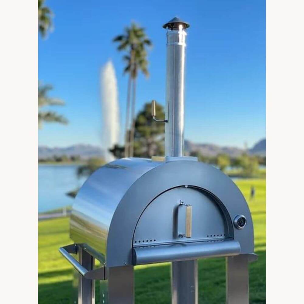 Close-up of the Kokomo 32” Outdoor Wood-Fired Stainless Steel Pizza Oven & Stand in a backyard, with tall palms, a fountain, and a clear blue sky in the background.