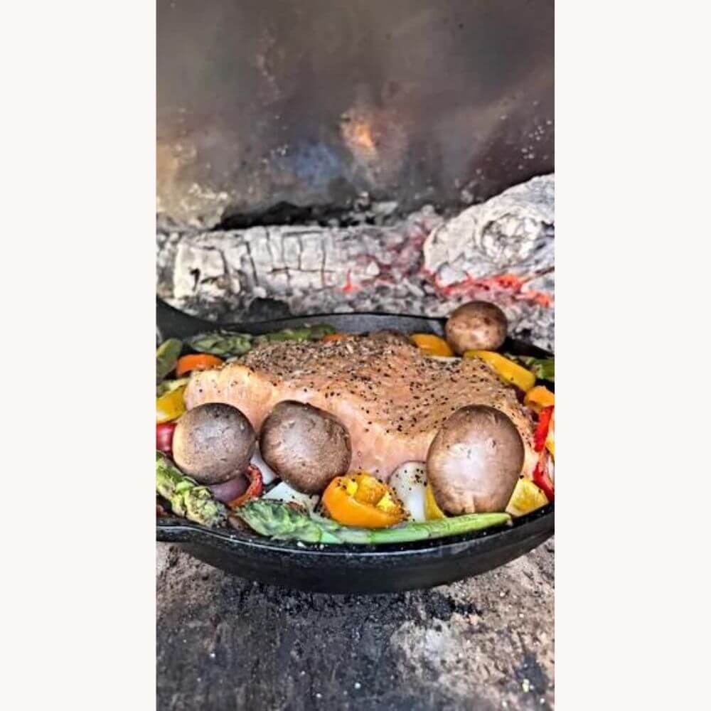 Salmon fillet seasoned with pepper, placed on a bed of colorful vegetables like mushrooms, bell peppers, and zucchini, in a black pan inside the Kokomo 32” Outdoor Wood-Fired Stainless Steel Pizza Oven.