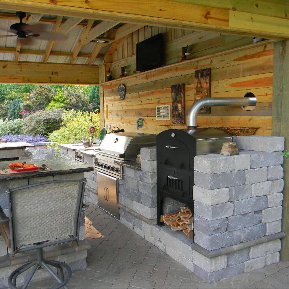 Tuscan Chef Medium Built-In Deluxe Family Pizza Oven (Model GX-CM) without cart, built into a firebrick countertop alongside a stainless steel grill and seating area under a wooden pergola, with greenery in the background.