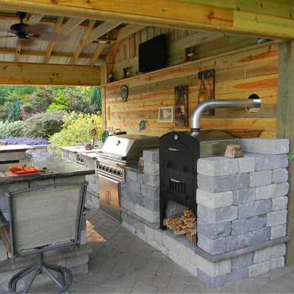 Tuscan Chef Medium Built-In Deluxe Family Pizza Oven (Model GX-CM) without cart, built into a firebrick countertop alongside a stainless steel grill and seating area under a wooden pergola, with greenery in the background.