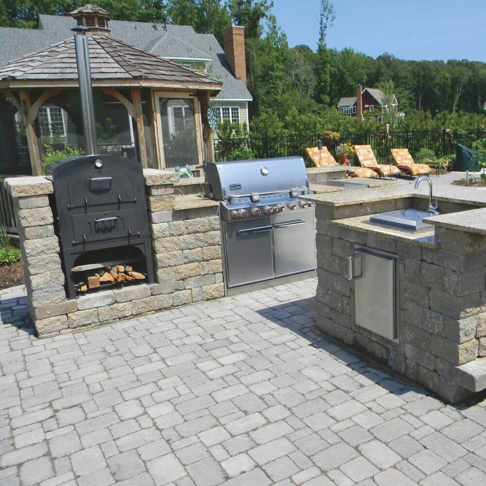 Tuscan Chef Medium Built-In Deluxe Family Pizza Oven (Model GX-CM) without cart, built into a stylish outdoor kitchen with a gas grill and sink on a paved patio, with a gazebo and lounge chairs in the background.