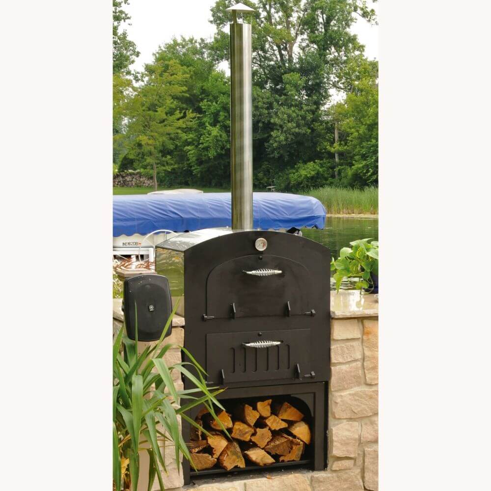 Tuscan Chef Medium Built-In Deluxe Family Pizza Oven (Model GX-CM) without cart, a black pizza oven with a tall chimney on a stone patio, with firewood stored below. Trees and a boat are in the background.