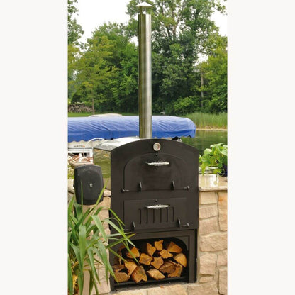 Tuscan Chef Medium Built-In Deluxe Family Pizza Oven (Model GX-CM) without cart, a black pizza oven with a tall chimney on a stone patio, with firewood stored below. Trees and a boat are in the background.