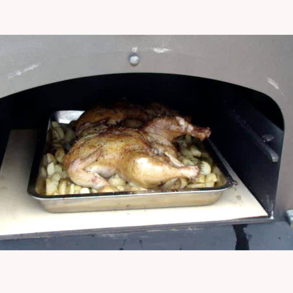 A whole roasted chicken sits on a bed of sliced potatoes in a metal baking tray inside a Tuscan Chef wood-fired pizza oven.