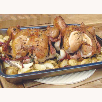 Two roasted chickens in a baking tray surrounded by roasted potatoes and onions, placed on a wooden surface.